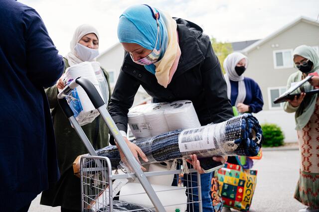 A woman garbed in a face mask and hijab bends over to grab donated items, including a rug and toilet paper, from a cart. Both are centered in the photo surrounded by four similarly garbed women helping to distribute donated items in support of the IRC.