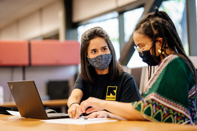 Carolina in an International Rescue Committee shirt and a woman in a green patterned dress, both masked, sit in front of a laptop at a table completing paperwork.