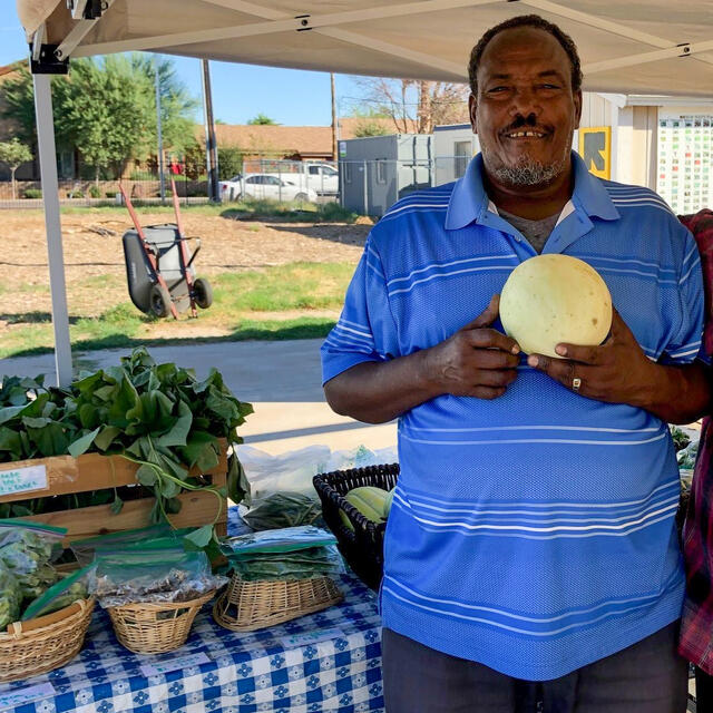 A man stands holding a vegetable next to a table of produce.