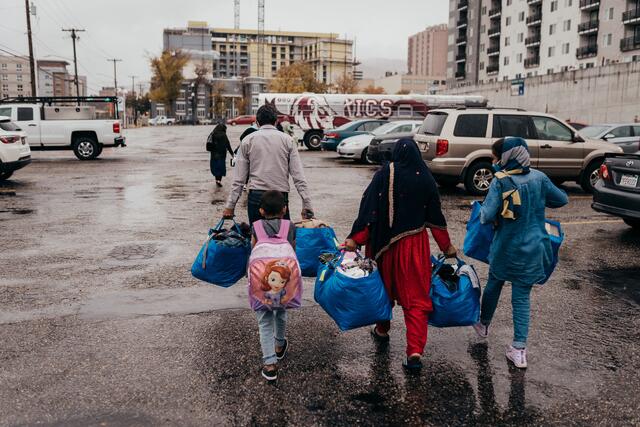 A family of four, with their backs facing the camera is walking in a parking lot toward a bus. They are carrying blue IKEA bags full of clothing.
