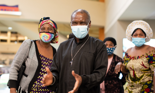 A family welcomes their daughter at the Boise airport after being apart for five years. Four family members, all masked, all smile as they see their daughter for the first time in five years. The father stands in the middle with his hands held out,