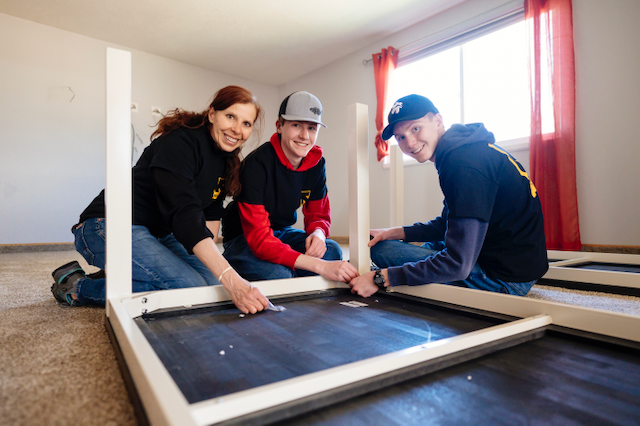 A mother and her two sons volunteering, building a white, metal frame table on the ground inside an apartment.