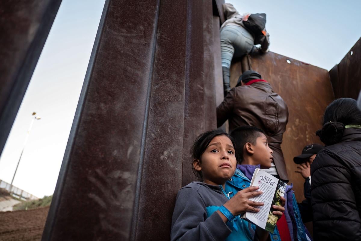 A Central American girl holds a book as others traveling in a caravan climb the Mexico-U.S. border fence in an attempt to cross to San Diego County.