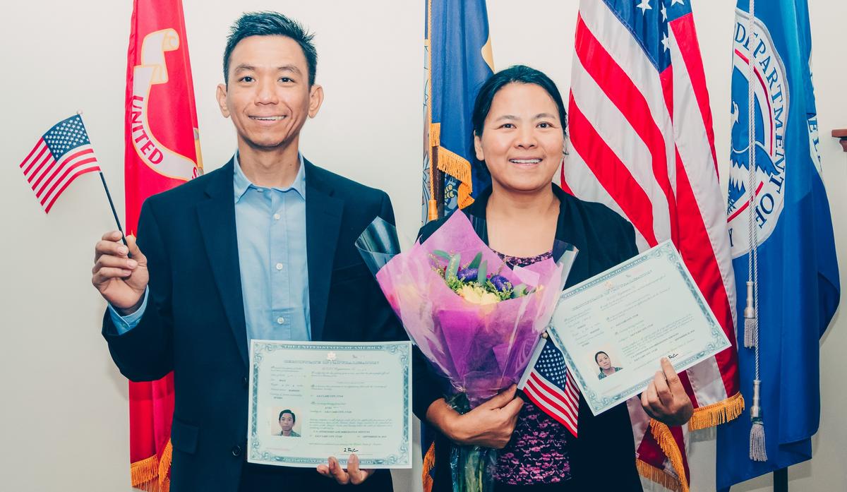 Sui and her husband Aung smile for the camera in front of large Utah and United States flags, holding small American flags, flowers, and their new naturalization papers.