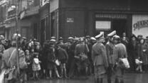 Ein schwarz-weiß Bild. Eine Gruppe Menschen steht vor einem Geschäft
