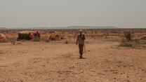 A man carrying a staff walks through a parched landscape in Ethiopia amid makeshift tents with mountains in the distance under a hazy sky.