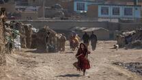 A girl in a red dress runs through a camp for displaced people in Kabul, Afghanista. There are people an makeshift homes in the background. 