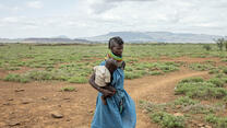 A Kenyan mother carries her baby across dry grassland with mountains in the background.