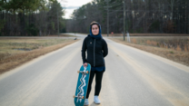 A 22 year old woman stands in the middle of a road holding a skateboard