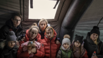 Eight Ukrainian refugees gather in a tent at the Medyka border crossing point in Poland. They all wear warm winter jackets.