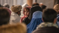 A mother holds a baby in the midst of a crowd of people. They wait to receive IRC cash assistance.