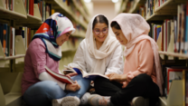 아리파, 자흐라, 하디사는 도서관 한가운데에 원을 그려 함께 앉아있습니다. 함께 책을 읽으며 웃고 있습니다.