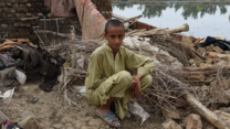A boy sits looking meekly at the camera. He sits in front of a pile of debris; the remnants of his home which was destroyed by flooding.