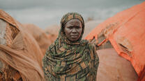 Portrait of a woman standing in front of a series of huts in the Somali desert.