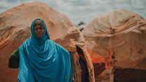 A woman poses for a photo outside of a small structure covered in blankets in Torotorow IDP Camp, Somalia.