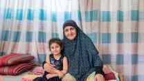 A young girl and her grandmother sit at the edge of a bed and pose for a photo.