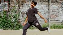 Ein junger Mann in einem schwarzen „buntkicktgut“-Trainingsanzug kickt einen Fußball auf einem Sportplatz vor einer Graffiti-bedeckten Wand.