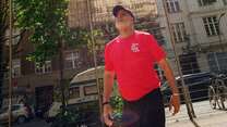 Ein Mann in einem roten T-Shirt und schwarzer Kappe schaut bei hellem Sonnenschein in die Luft, während ein Ball über ihm schwebt; im Hintergrund sind ein Zaun und mehrstöckige Gebäude zu sehen.