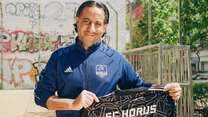 Ein Mann im SC Horus Trainingsanzug hält stolz ein Trikot mit der Nummer 1 vor einem mit Graffiti bedeckten Gebäude hoch.