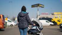 Eine Person, die einen Kinderwagen durch einen Parkplatz schiebt, umgeben von verschiedenen Fahrzeugen und Gebäuden bei klarem Himmel.