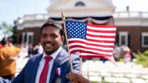 A man stands in front of a building, smiling and posing for a photo while holding the U.S. flag.