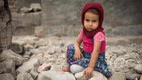 Small girl sits among rubble looking at the camera.