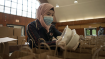 Rania Abou, wearing a mask, puts two containers of food inside paper bags to be given away as part of her work with the IRC and World Central Kitchen.