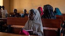 Young girl Fatima sits in a class room looking up towards where the teacher stands. 