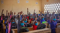 A classroom full of school children raise their hands.