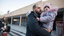 A Ukrainian refugee man in black coat holds his child in silver coat with pink hat