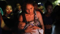 어둠 속에서 미국-멕시코 국경으로 데려가 줄 버스를 기다리는 중미 망명 신청자 속에 아기를 안고 있는 어머니가 서 있습니다.