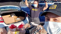 Two IRC staff members wearing IRC shirts and hats stand next to a car with an open trunk. In the trunk are groceries they recently bought for refugee and immigrant families. 