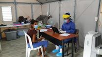 A woman sits at a chair at a table with doctor in full PPE opposite her. They are in a temporary clinic set up by the IRC at the Simón Bolívar bridge. 