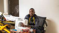 Jonathan Amissa sits at a desk with a sewing machine he uses to make masks for his staff and community. He is wearing yellow tape measure around his neck and is looking at the camera and smiling. There is a stack of red masks on the table.  