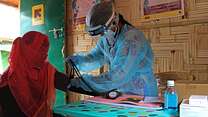 An IRC midwife in Bangladesh, wearing a mask, face shield and protective suit, reads the blood pressure of a patient whose face is covered by a veil to guard against COVID-19.