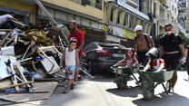 Two men push children in wheelbarrows past damage from the Aug 4, 2020 explosion in Beirut, Lebanon. Getty photo