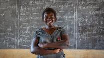 Jackie Letaru, a teacher and activist in Uganda, poses smiling in front of a chalkboard 