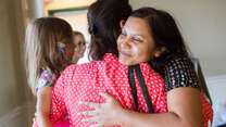 Two women hug while one holds a small child. We see one of their faces, she is smiling. 