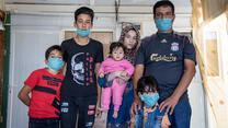 In a small tin home in a refugee camp, Manhal poses with his family: his wife holds a baby in a pink onesie, his four-year-old stands in front of him and eight-year-old and fifteen-year-old sons stands next to them. 