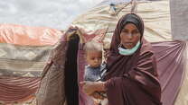 Amina holds her son while standing in front of several tents. 