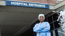 Nabila stands with her arms crossed and smiling in front of the hospital where she works. She is wearing blue scrubs and a white winter hat. 