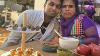 Sitting at a table with an array of food in front of them, Shanthini and Sarujen lean toward one another and look at the camera. 