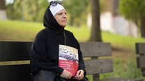 Maha al-Obaidi sits on a park bench looking off into the distance and holding a U.S. citizenship test study book. 