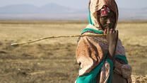 A teenage girl, holding a stick and looking at the camera, stands in a drought affected very dry landscape with mountains in the distance. 