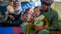 A mother and father in Afghanistan sit on the ground among blankets and pillows, each with a child on their lap. The boy in the front is smiling and his father is looking at him. 