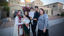 Muska, a woman in her twenties, stands in front of her home with three adult family members, a toddler and a baby. 