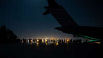 A night shot, with the tail of an airplane in the foreground and a line of people in the background. We can see a light in the distance and just the silhouette of the plane and people. 