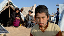 A boy looks into the camera as two elderly people sit on sandy ground behind him in front of their tent 