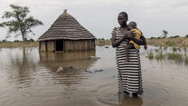 With water above her ankles, a mother holds her child and stands in front of her flooded home. 