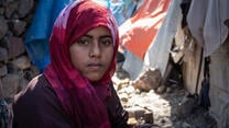 11살 먹은 소녀가 예멘 국내 실향민 캠프 밖에 앉아서 카메라를 응시합니다. 이 소녀의 뒤에는 빨래가 걸려 있습니다. 